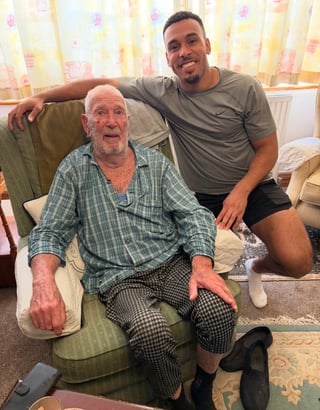 A younger man in a gray shirt stands beside an elderly man in a plaid shirt, both smiling at the camera in a home setting.